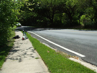 We are looking to the left along a two-lane street with curb, grass and sidewalk on the left and thick trees on the other side of the sidewalk.  On the right side are thick trees starting at the edge of the street.  The road is going slightly downhill and curves gently to the left, disappearing behind the trees about 100 feet away.